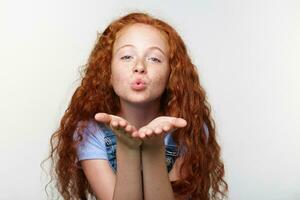 Portrait of cheerful little girl with ginger hair and freckles, send kiss at the cam, looks happy, standing over white wall. photo