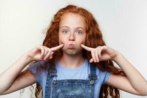 Joyful nice little girl with ginger hair and freckles, looks at the camera, blowing cheeks and lips, touches cheeks with fingers and looks funny, stands over white background. photo