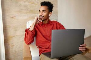 Positive young attractive bearded dark skinned male with short haircut touching his face with raised hand and smiling slightly while looking at window, working remotely from home photo