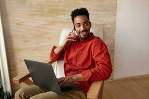 Cheerful young lovely short haired bearded male with dark skin smiling widely aside while making call, working remotely from home, isolated over beige background photo