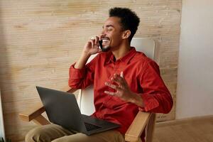 Side view of good looking young cheerful short haired bearded male with dark skin keeping his eyes closed while having pleasant talk, sitting in chair over home interior photo