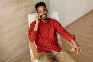 Happy young attractive brunette bearded dark skinned guy dressed in casual wear looking gladly at camera with pleasant smile while having phone talk, isolated over home interior photo