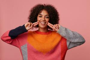 Happy attractive young curly dark skinned brunette lady with casual hairstyle keeping fingers on her cheeks while smiling widely, wearing multi-colored woolen sweater over pink background photo