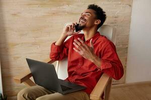 Overjoyed young pretty short haired bearded dark skinned man thowing back his head while laughing and raising emotionally hand, making phone call while sitting in chair photo