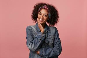 Cheerful lovely curly brunette female with dark skin wearing casual hairstyle, keeping raised hand on her cheek while smiling happily to camera, isolated over pink background photo