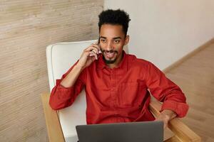 Cheerful young pretty short haired dark skinned brunette male dressed in red shirt smiling gladly while having pleasant phone conversation, sitting over home interior photo