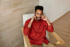 Indoor shot of young brown-eyed short haired bearded male with dark skin leaning his face on raised hand and looking confusedly at camera while making call photo