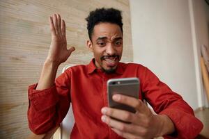 Indoor photo of bewildered young bearded dark skinned male looking excitedly at screen of his mobile phone and keeping hand raised, isolated over home interior