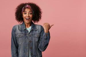 Studio photo of happy young lovely dark skinned female with short curly brown hair posing over pink background in colorful headband and jeans coat, looking cheerfully to camera with excited face