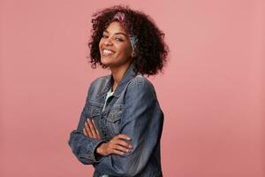 Studio shot of young charming dark skinned woman in colorful headband having short curly hair, smiling happily to camera while posing over pink background with folded hands photo