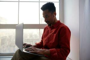 Indoor photo of young bearded dark skinned brunette guy preparing materials for meeting and typing text on his laptop, posing on windowsill in casual clothes