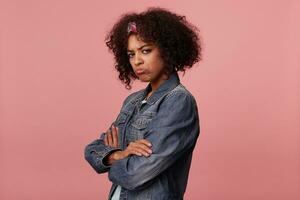 Offended young dark skinned curly brunette female with short haircut wearing colorful headband and twisting her lips while looking to camera, dressed in casual wear over pink background photo