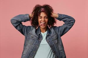 Portrait of cheerful dark skinned curly brunette lady in colorful headband keeping raised hand on her head while winking joyfully to camera and showing her tongue, isolated over pink background photo