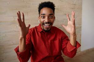 Agitated young brown-eyed short haired curly bearded guy raising emotionally his hands while looking joyfully at camera with broad smile, isolated over beige interior photo