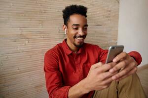 Glad young handsome short haired dark skinned curly guy holding mobile phone and smiling happily while reading messages, posing over beige interior in red shirt photo