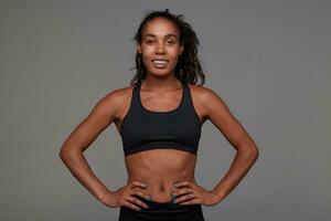 Pleasant looking young dark skinned curly brunette female in good physical shape looking positively at camera and smiling slightly, standing against grey background photo