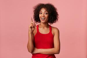 Studio photo of young smart African American lady wearing a red t-shirt, with curly dark hair. Smiling, shows index finger up, because have a great idea. Isolated over pink background.