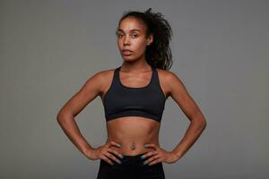 Studio shot of young pretty dark skinned curly lady with belly button piercing holding hands on her waist while posing over grey background in athletic black clothes photo