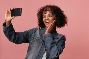 Studio photo of attractive positive young curly brunette woman with dark skin smiling widely and keeping palm on her cheek while making selfie on her smartphone over pink background