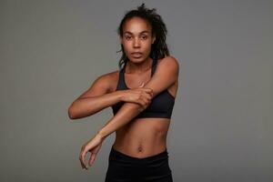 Indoor photo of young dark skinned curly lady with casual hairstyle preparing for evening workout after working day, standing against grey background in sporty wear