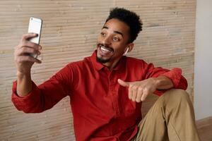 Happy young handsome short haired bearded brunette man smiling happily while having video call with his mobile, sitting over beige interior in casual clothes photo