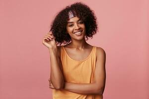 Portrait of cheerful young curly brunette female with dark skin pulling her hair while looking to camera with wide sincere smile, wearing light orange shirt over pink background photo