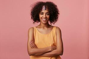 Portrait of cheerful attractive young dark skinned lady with short curly brown hair looking positively at camera with pleasant smile, folding hands on her chest while standing over pink background photo