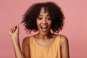 Joyful brown-eyed lovely dark skinned female with short brown hair pulling her curls and looking cheerfully at camera with wide mouth opened, isolated over pink background in colorful casual clothes photo