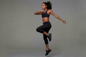 Full-length shot of slim young dark skinned curly brunette female being concentrated while making physical exercises in fitness studio, standing against grey background photo