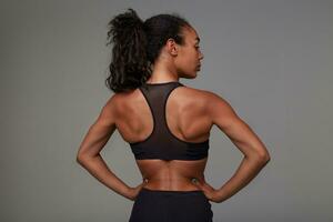 Back view of young sporty dark skinned curly woman with ponytail hairstyle holding her hands on waist and demonstrating her perfect body, standing against grey background photo