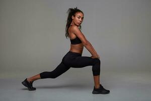 Fitness young attractive dark skinned curly model stretching her legs and looking at camera with folded lips, wanting to be fit and healthy, posing over grey background photo