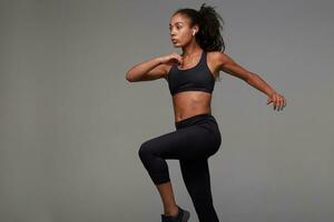 Shot of young pretty curly brunette woman with dark skin training indoor in black clothes, listening to music with earphones while posing over grey background photo