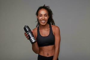 Cheerful young attractive curly brunette female with dark skin being in nice mood while standing over grey background, smiling widely with fitness bottle in raised hand photo