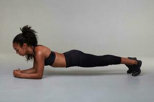 Full-length photo of young athletic dark skinned curly female with good body doing plank exercise indoor, standing over grey background in black top and leggins. Sport and wellness concept