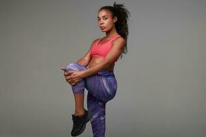 Studio photo of young sporty dark skinned woman wearing her curly long brown hair in ponytail hairstyle while stretching her muscules after hard training, isolated over grey background