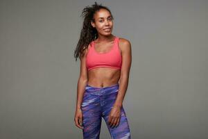 Attractive young dark skinned curly brunette woman dressed in pink sporty top and printed leggins, looking positively at camera with sincere smile, standing against grey background photo