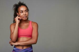 Pensive athletic young dark skinned curly lady with casual hairstyle looking thoughtfully aside and touching her face gently, dressed in sporty wear over grey background photo