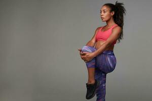 Fitness young dark skinned curly model with ponytail hairstyle posing over grey background in sporty pink top and printed leggins, stretching muscules of her legs photo