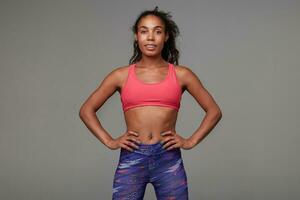 Positive active young dark skinned curly brunette woman with belly button piercing looking cheerfully at camera while standing over grey background. Fitness and sport concept photo