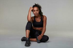 Indoor photo of young charming dark skinned woman with long brown curly hair making break with workout and drinking water from fitness bottle, posing over grey background