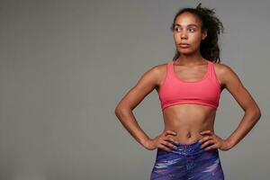 Studio shot of young attractive dark skinned curly woman with ponytail hairstyle looking aside with folded lips and keeping her hands on waist, isolated over grey background photo