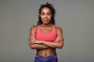 Positive young slim curly brunette woman with dark skin folding hands on her chest while smiling pleasantly at camera, being in good physical shape, standing against grey background photo