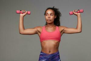 Fitness young slim dark skinned curly brunette female looking aside with folded lips while making exersices with dumbbells, goes in for sport every morning, isolated over grey background photo