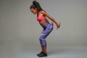 Side view of sporty young dark skinned curly long haired woman dressed in athletic clothes holding dumbbells behind her back while making exercises for hands, isolated over grey background photo