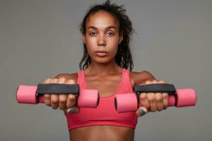 Portrait of sporty young brown-eyed dark skinned curly lady holding weighting agents in raised hands and looking at amera with calm face, wearing athletic pink top over grey background photo