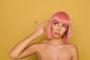 Studio shot of young attractive woman with short pink hair folding gun with raised hand and keeping it on her temple while posing over mustard background with naked shoulders photo