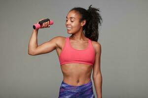 Positive sporty young dark skinned curly lady with ponytail hairstyle looking happily on her raised hand, being proud of her strong biceps, isolated over grey background photo