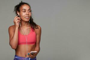 Studio shot of young slim curly long haired brunette lady with dark skin inserting earpiece into her ear and looking aside with pleasant smile, relaxing after gym over grey background photo