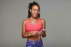 agradable mirando joven aptitud oscuro desollado Rizado modelo con casual peinado disfrutando música pista con auriculares y móvil teléfono mientras en pie terminado gris antecedentes foto