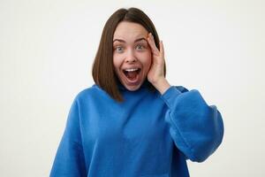 Amazed young lovely brown haired lady with short haircut raising hand to her face while looking dazedly at camera with wide mouth opened, posing over white background photo